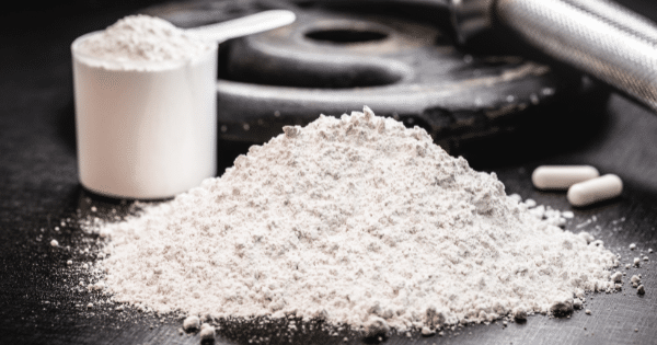 A scoop and pile of white supplement powder next to two capsules and a weight plate on a dark surface.