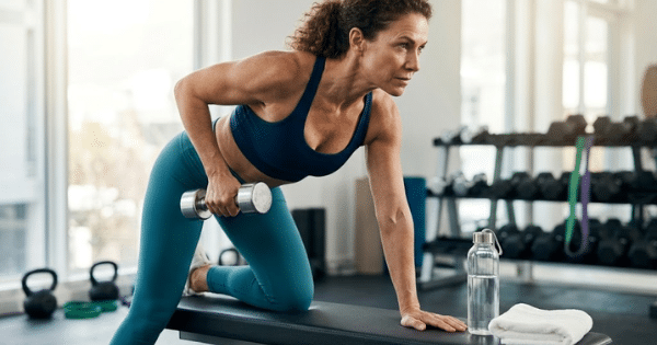 A woman in workout attire performing a one-arm dumbbell row with one knee and hand resting on a workout bench.