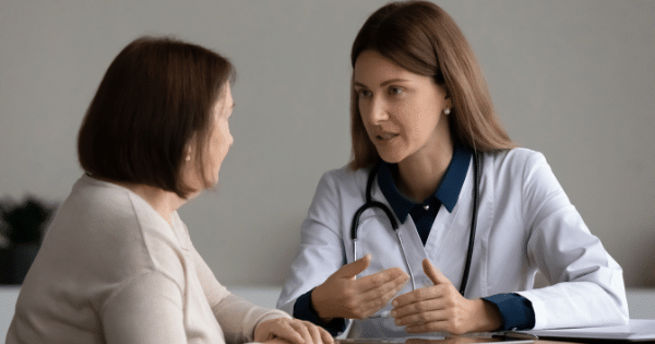 A female doctor in a white lab coat talking attentively to a patient across a table.