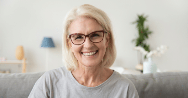 A headshot of a smiling senior woman with blonde hair and eyeglasses sitting on a couch.