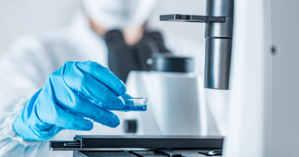 A gloved laboratory worker placing a petri dish containing a blue liquid under a microscope.