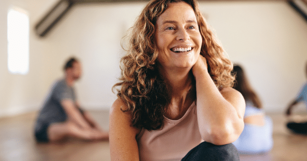 A smiling middle-aged woman with curly hair resting her head on her hand during a group yoga or workout session.