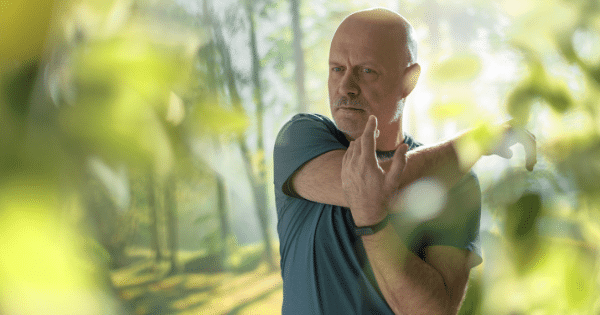 A mature man stretching his arm across his chest outdoors in a sunlit park setting.