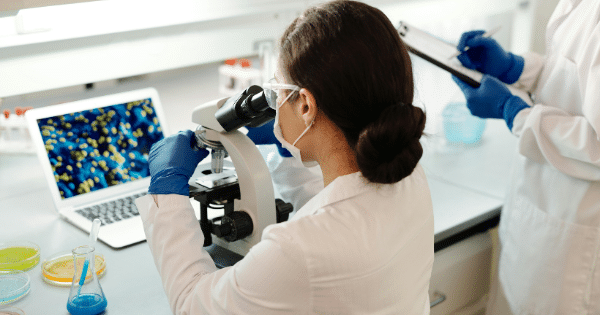 A female researcher using a microscope while a colleague takes notes on a clipboard in a laboratory