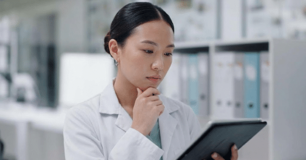 An Asian female researcher thoughtfully reviewing data on a tablet in a laboratory or office setting.