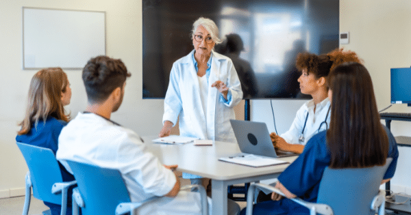 A senior female instructor or doctor speaking to a group of medical professionals seated around a conference table.