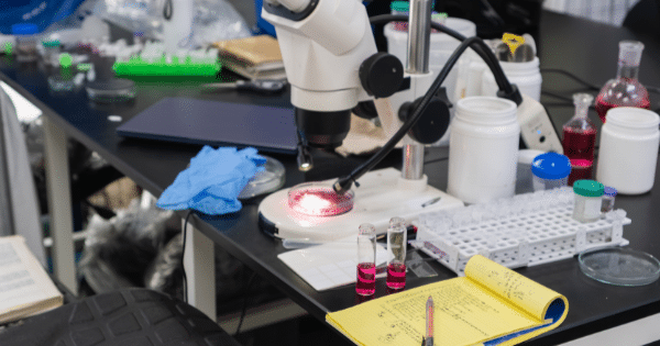 A cluttered laboratory workbench featuring a stereo microscope, liquid vials, petri dishes, and a yellow notepad.