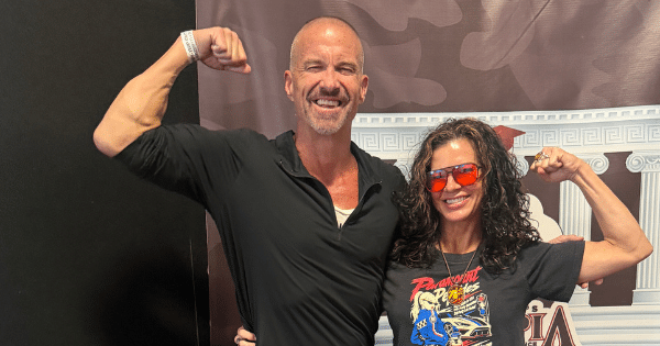 A smiling man and woman standing together while flexing their biceps in front of an event backdrop.