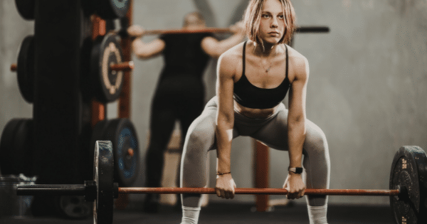 A athletic woman in a gym preparing to perform a sumo deadlift, gripping a loaded barbell while maintaining a wide squat stance.