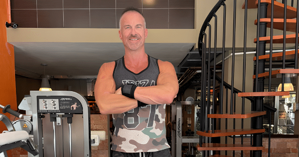 A fit, smiling man in a camouflage gym tank top standing with his arms crossed in front of exercise equipment and a spiral staircase in a fitness center.