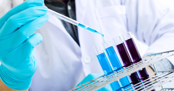 A researcher wearing blue protective gloves uses a pipette to add liquid into test tubes held in a laboratory rack.
