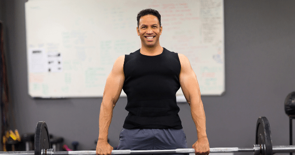 A smiling man in athletic attire standing in a gym holding a barbell loaded with weight plates.