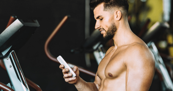 A side view of a shirtless, fit man looking down at his smartphone while standing near exercise equipment in a gym setting.