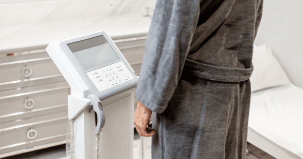 A person wearing a plush gray bathrobe stands next to a white body composition analyzer machine in a room with a examination bed.