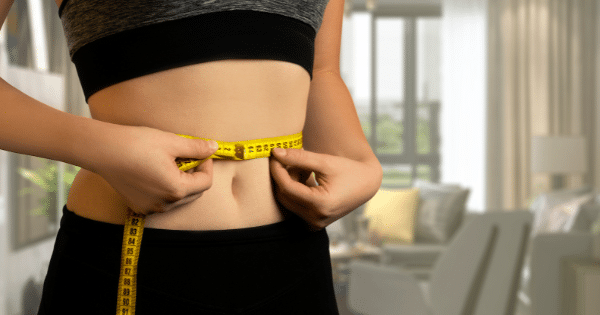 A close-up shot of a person using a yellow measuring tape wrapped around their waist in a living room setting.