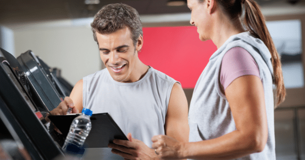A personal trainer in a gray tank top holding a clipboard and smiling while reviewing notes with a female client at a gym.