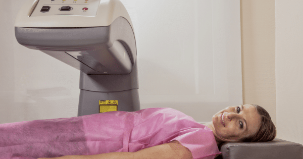 A woman in a pink gown lying down comfortably on a DEXA bone density scan bed, smiling toward the camera.
