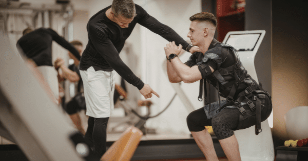 A trainer guides a man wearing an EMS (Electrical Muscle Stimulation) suit as he performs a squat in a gym.