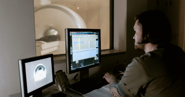 A medical technician sitting at a computer workstation in a control room, monitoring scanning computer displays with a clear view into an MRI scanner room.