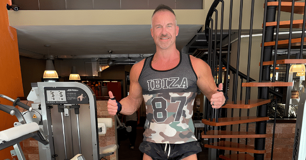 A fit man in a camouflage-and-black printed tank top giving two thumbs up while standing in a gym next to a spiral staircase and workout machines.