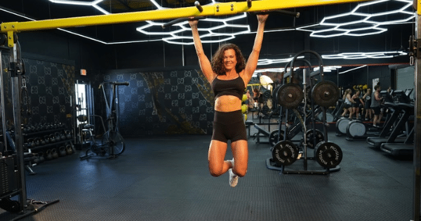 A fit woman in black athletic wear hangs from a yellow pull-up bar inside a modern gym with hexagon ceiling lights.