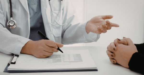 A doctor in a lab coat and scrubs holds a pen over medical documents while gesturing during a consultation with a patient seated across the table.