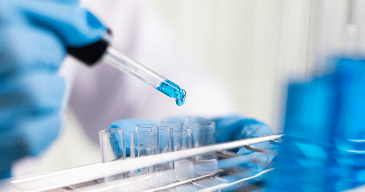 A gloved hand dispenses a drop of blue liquid from a pipette into laboratory test tubes.