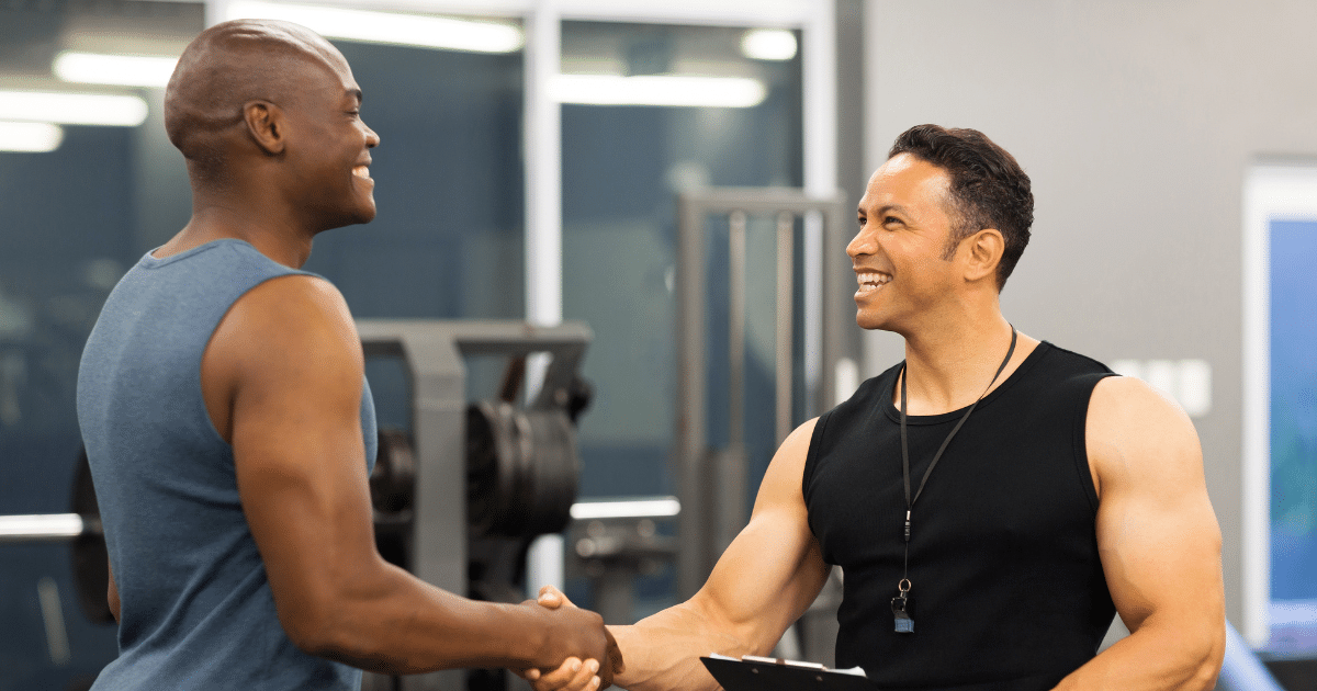 A smiling personal trainer shaking hands with a male client inside a gym.