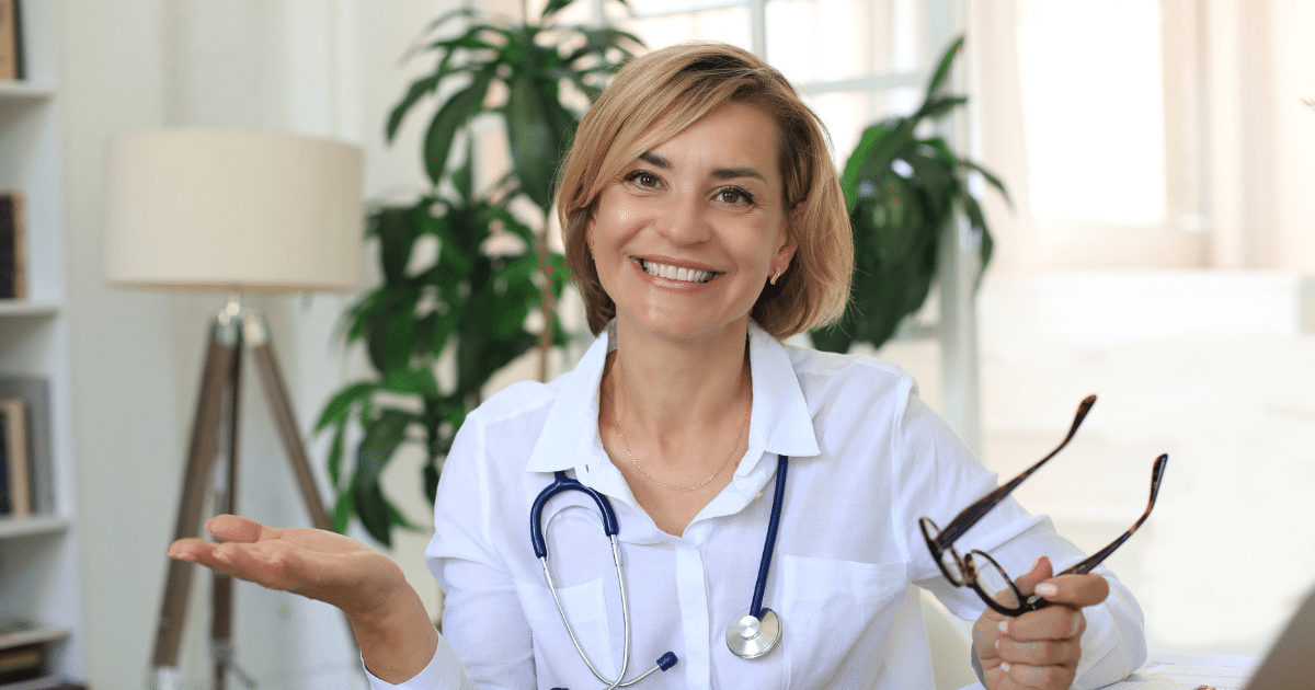 A smiling female wearing a stethoscope around her neck, holding eyeglasses in one hand and gesturing with the other.