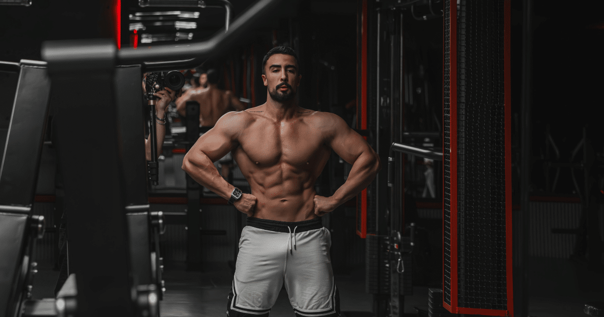 A muscular, shirtless man with a beard stands inside a gym, posing front-facing with his hands on his hips to show off his physique and abdominal muscles.