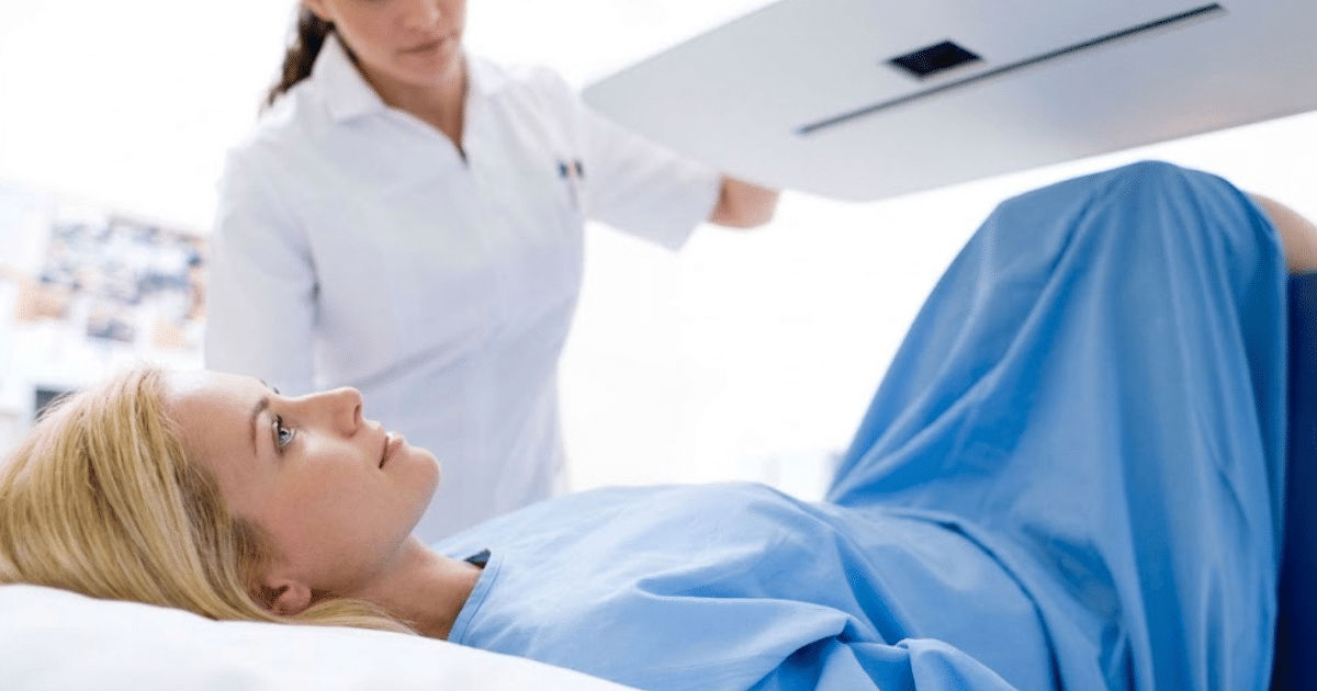 A blonde woman in a light blue hospital gown lies on a medical table looking up while a healthcare provider stands over her positioning an imaging scanner overhead.