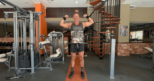 A muscular man flexing both biceps inside a gym in front of workout equipment and a spiral staircase.